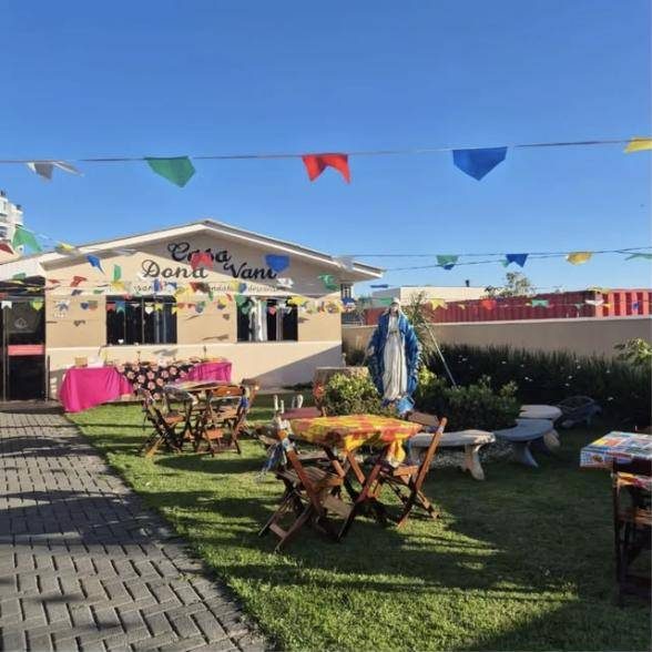 Quintal da Casa Dona Vani enfeitado com bandeirinhas, mesas e cadeiras no gramado, com a casa ao fundo